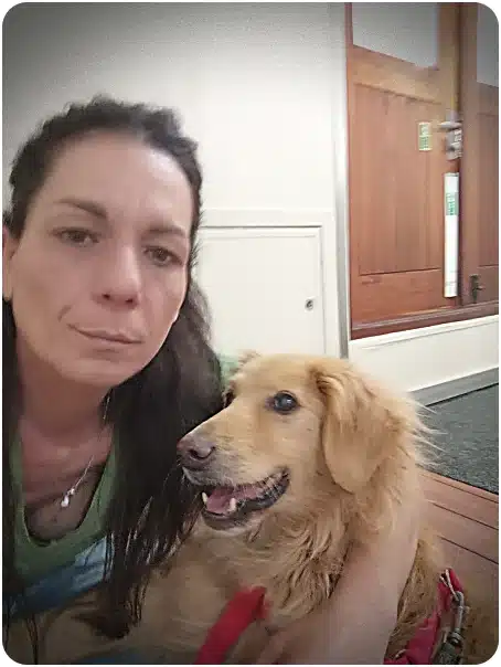 Woman with dark hair sits beside a golden retriever in a hallway, both looking at the camera.