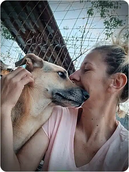 Woman in a light pink shirt cuddles and kisses a tan dog pressed up against a chain-link fence.
