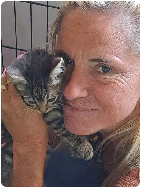 Smiling older woman with light hair holding a gray tabby kitten close to her cheek.