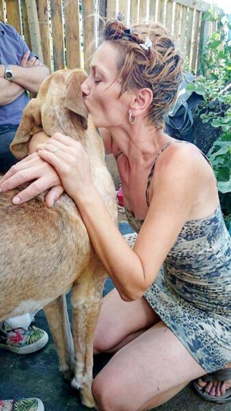 Woman kneels and kisses a light brown dog on the muzzle in a fenced yard, showing affection.