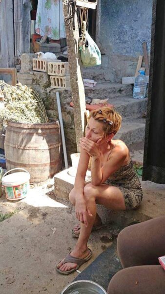 A woman in a patterned dress sits on concrete steps in a rustic outdoor area, resting her chin on her hand and wearing sandals.