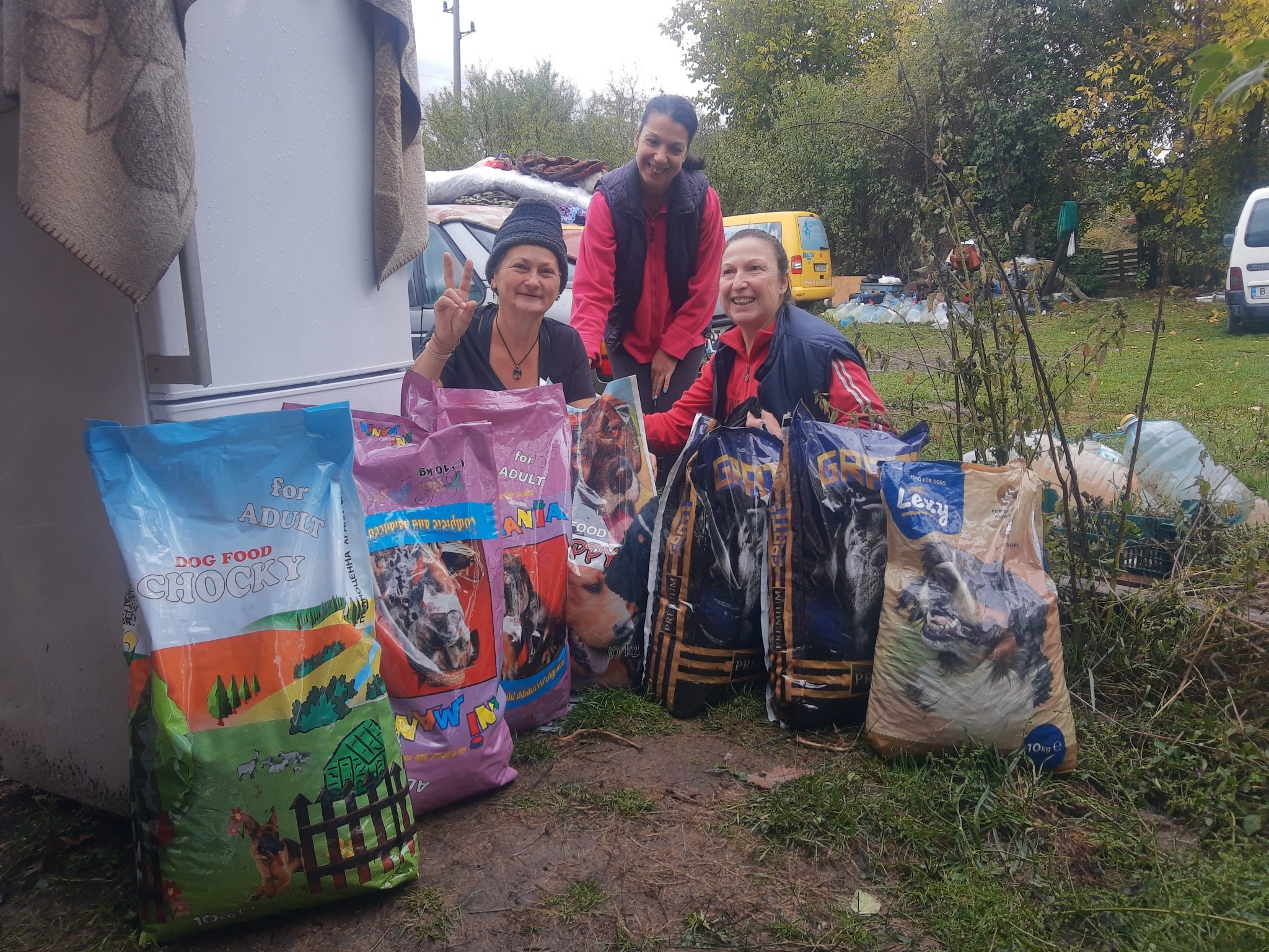 Three women smile among stacked dog-food bags outdoors, likely a donation or relief setup.