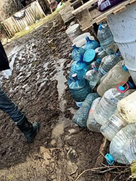Mud-covered ground beside a stack of water jugs and buckets near a wooden structure; a person in boots stands nearby.