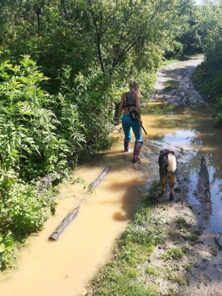 Woman wading through muddy puddles on a forest trail with a dog nearby, wearing blue pants and boots.