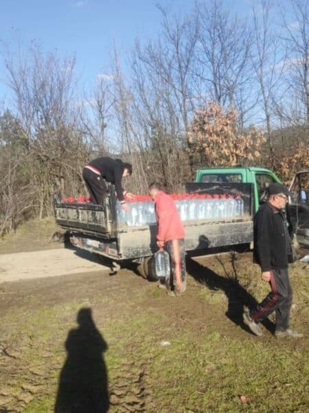 Two men loading large bottled water onto the back of a pickup truck in a rural area with bare trees in autumn or wintertime.