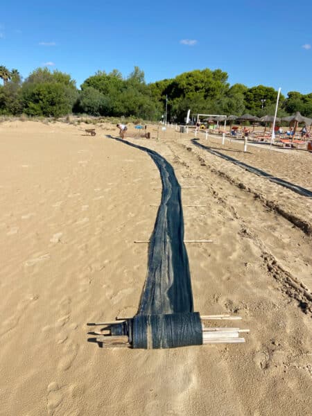 Black geotextile fabric unfurling along a sandy beach as workers prepare a barrier, with trees and beach chairs in the background.