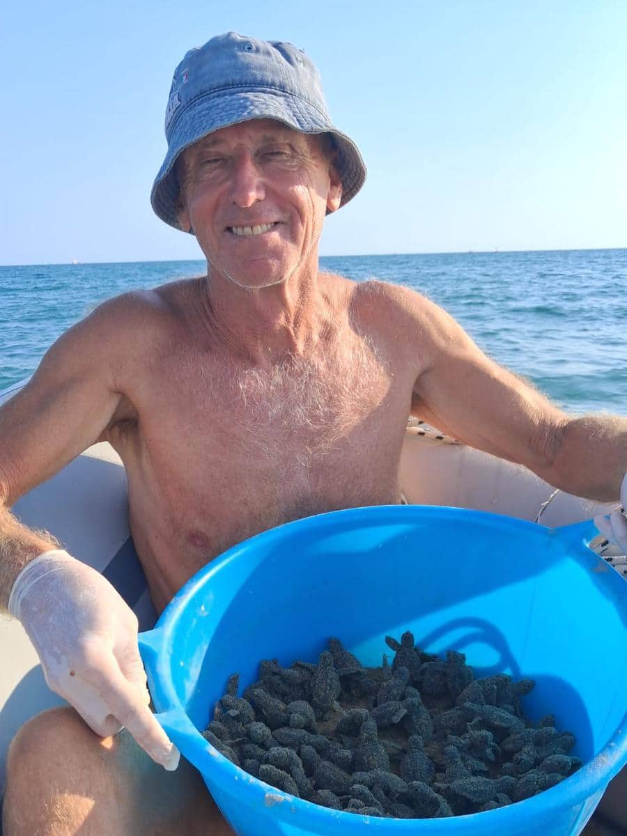 Older man wearing a blue bucket hat on a boat at the ocean, holding a blue bucket filled with small dark sea creatures or shellfish.