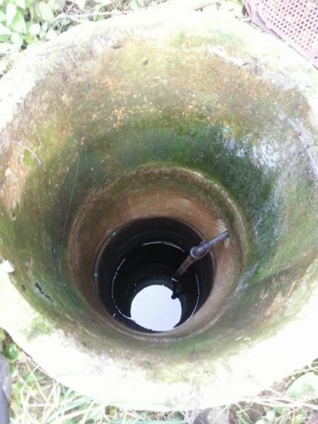 Open shaft or well with mossy, green walls, looking down to a dark circular interior with water at the bottom and a metal tool extending inside