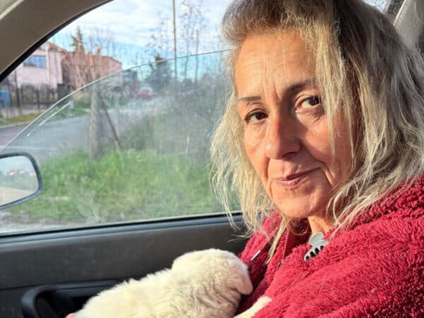 Woman sitting in a car with a small white dog asleep on her lap, wearing a red fleece jacket.