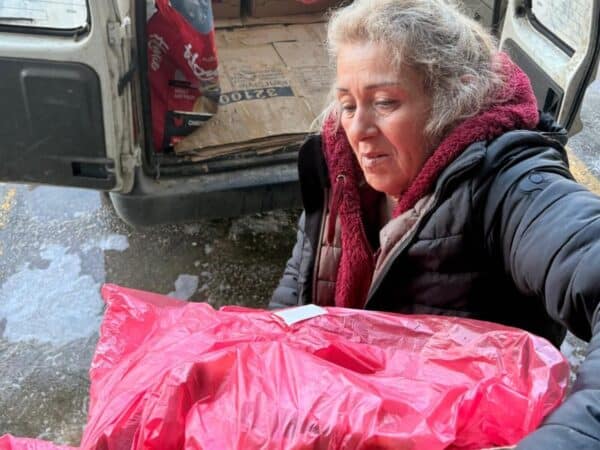 Older woman in a dark winter jacket and red scarf stands beside an open van loaded with cardboard; a pink plastic bag is in the foreground.