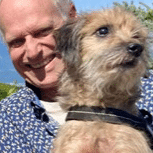 Smiling man in a blue patterned shirt with a small scruffy brown dog wearing a black harness outdoors, both facing the camera.