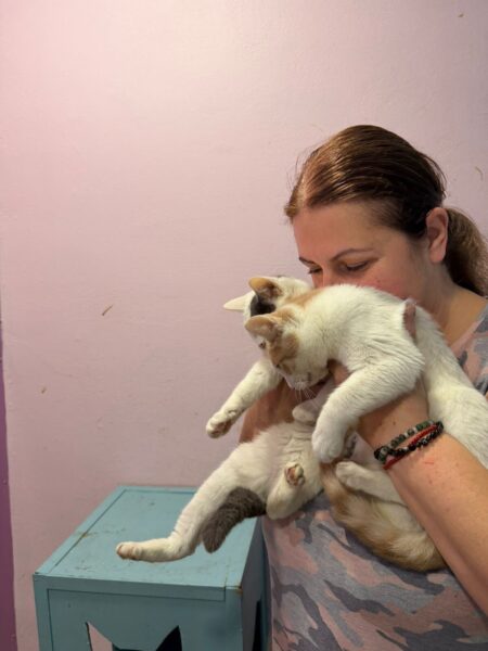 Woman holds a white and orange cat up to her chest, pink wall behind them as they cuddle closely.