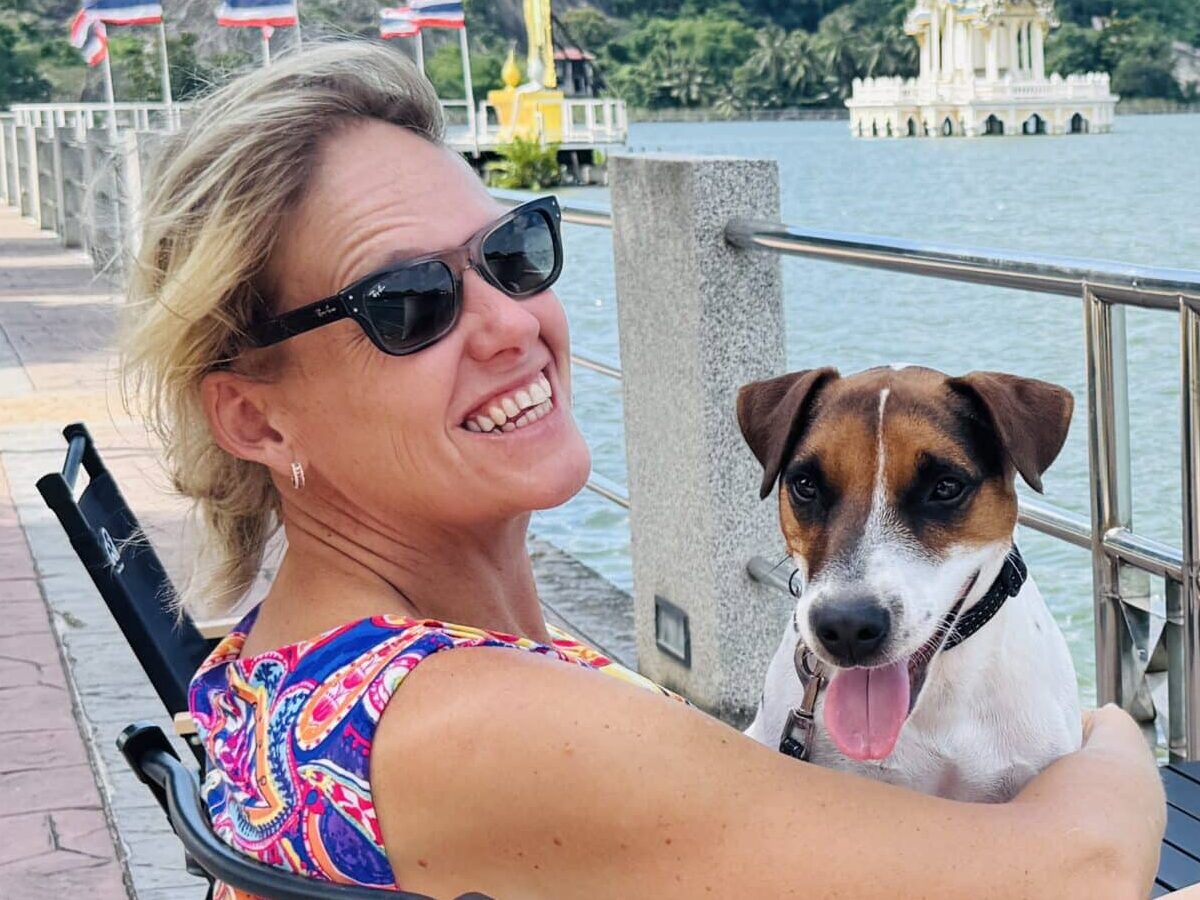 Smiling blonde woman in sunglasses sits with a brindle and white dog by a waterfront railing on a sunny day, colorful top visible.