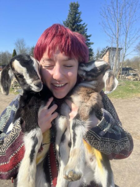 Person with short red hair smiling as two goats nuzzle against their cheeks outdoors on a sunny day.