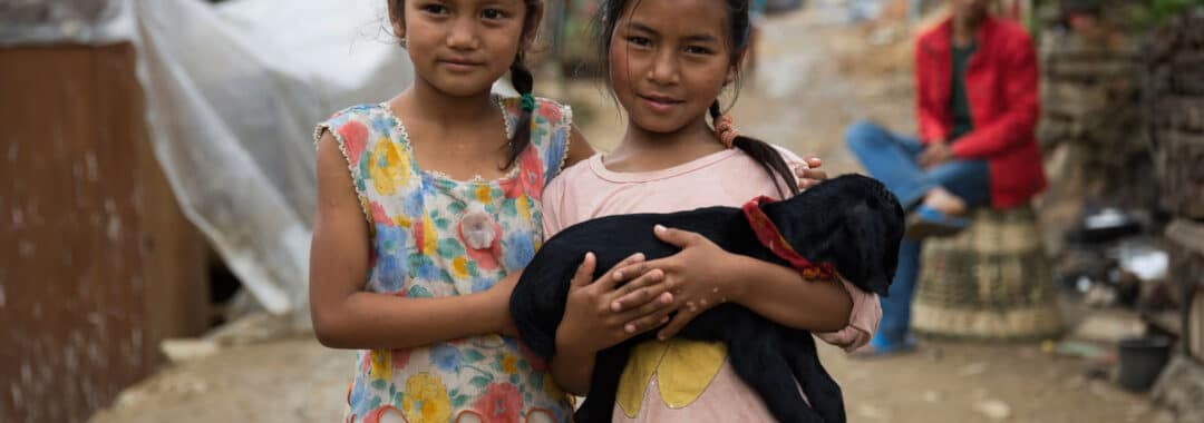 Two young girls stand side by side in a muddy village street, holding a black puppy together.