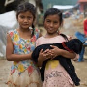 Two young girls stand side by side in a muddy village street, holding a black puppy together.