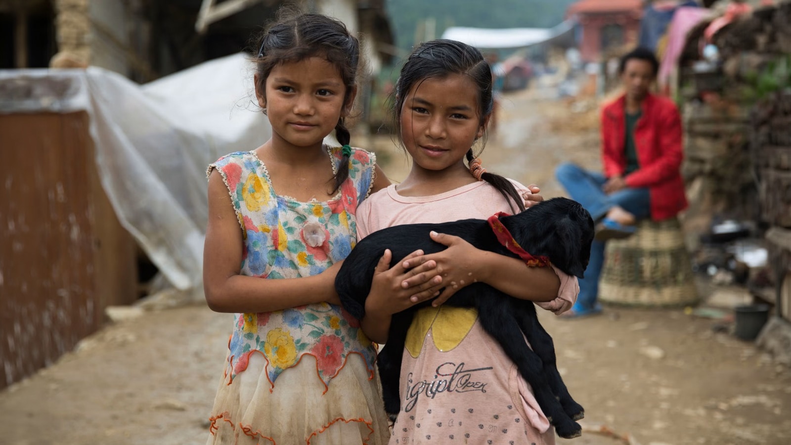 Two young girls stand side by side in a muddy village street, holding a black puppy together.