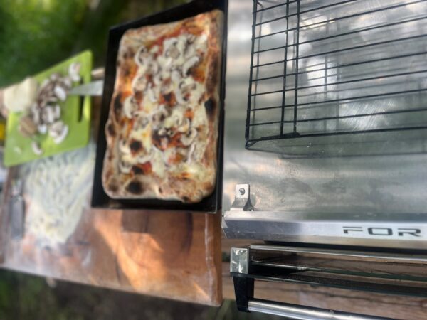 Pizza baking on a metal tray in a grill/oven, with a green cutting board and toppings nearby on the counter.