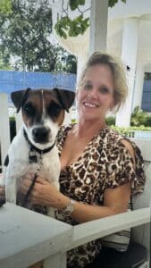 Smiling woman in a leopard-print blouse sits on a porch with a brown-and-white dog on a leash nearby.