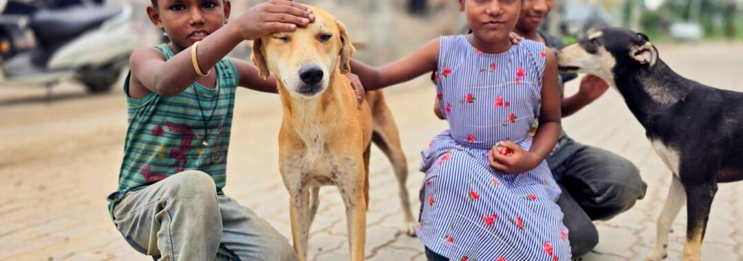 Three children kneel on a brick street, petting a tan dog between them; another dog stands nearby in the background.