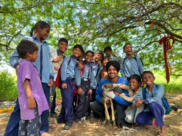 A group of smiling children and a man kneeling with a dog outdoors under a leafy tree.