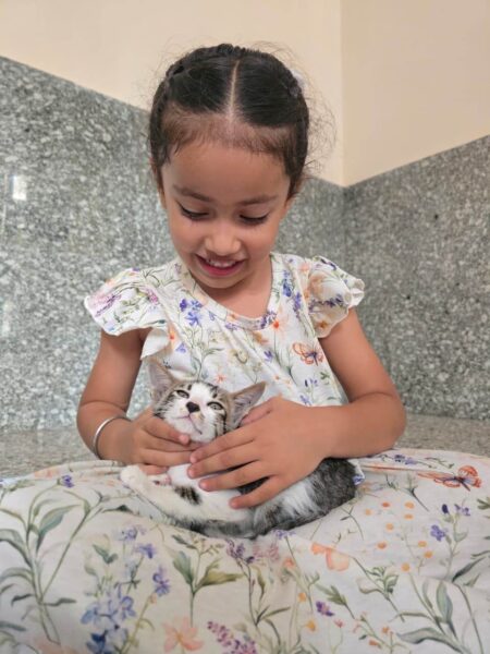 Young girl in a floral dress gently holding a kitten on her lap, both looking down with smiles in a tiled room.