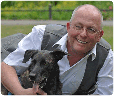 Smiling man with glasses posing with a black and gray dog outdoors in a park setting.